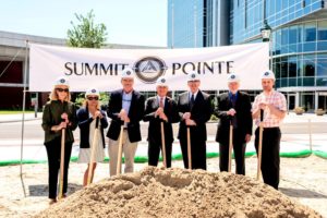 7 men and women stand around a dirt pile with shovels..